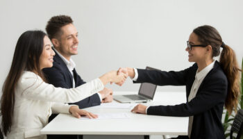Smiling diverse businesswomen shake hands at group meeting, friendly asian hr handshaking congratulating hired applicant at job interview, satisfied millennial partners make contract deal concept